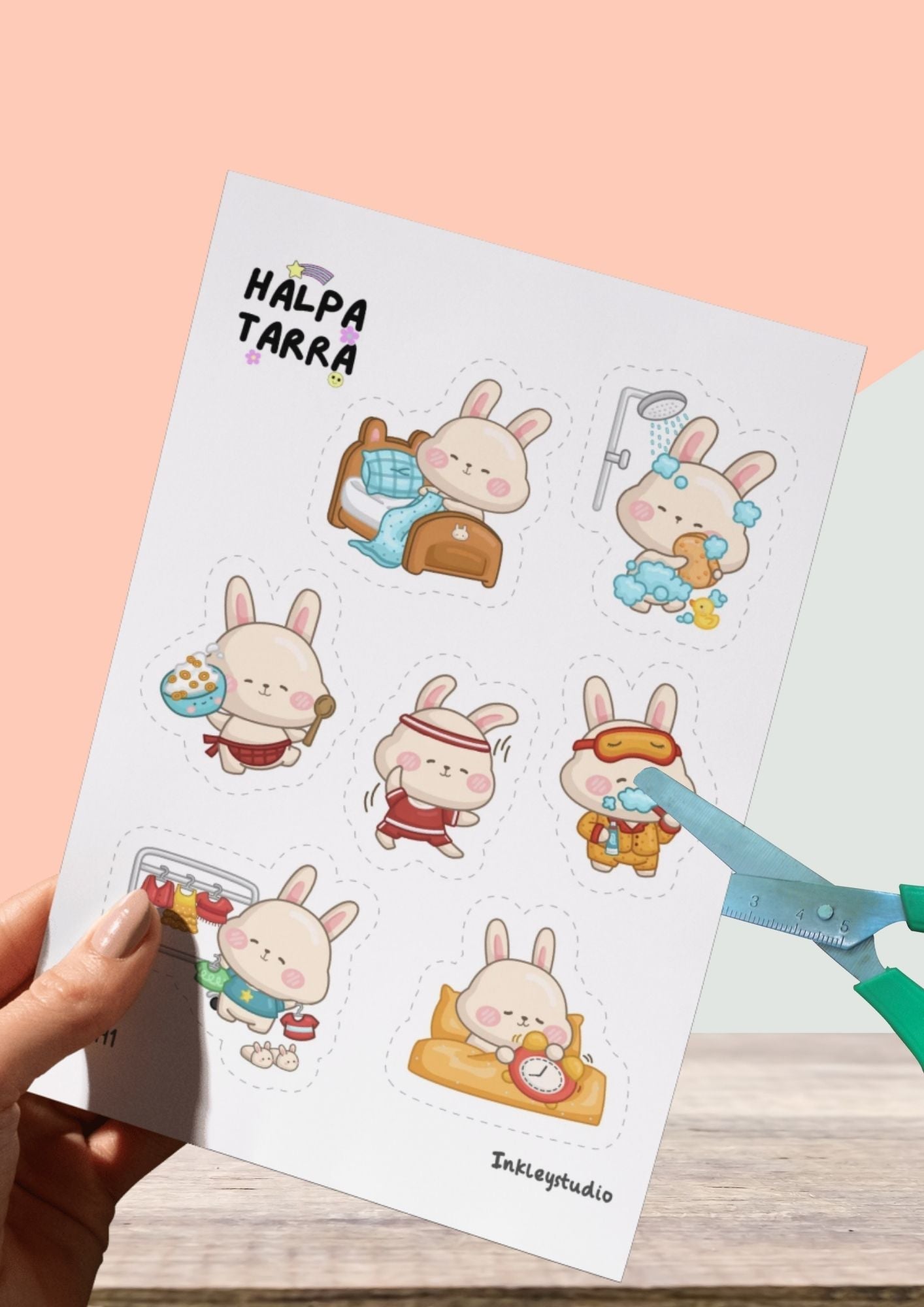 Käsi pitää leikkaa ja liimaa -tarra-arkkia, jossa on suloisia pupu-aiheisia kuvia katkoviivoilla. Vieressä saksilla leikataan tarraa katkoviivaa pitkin.